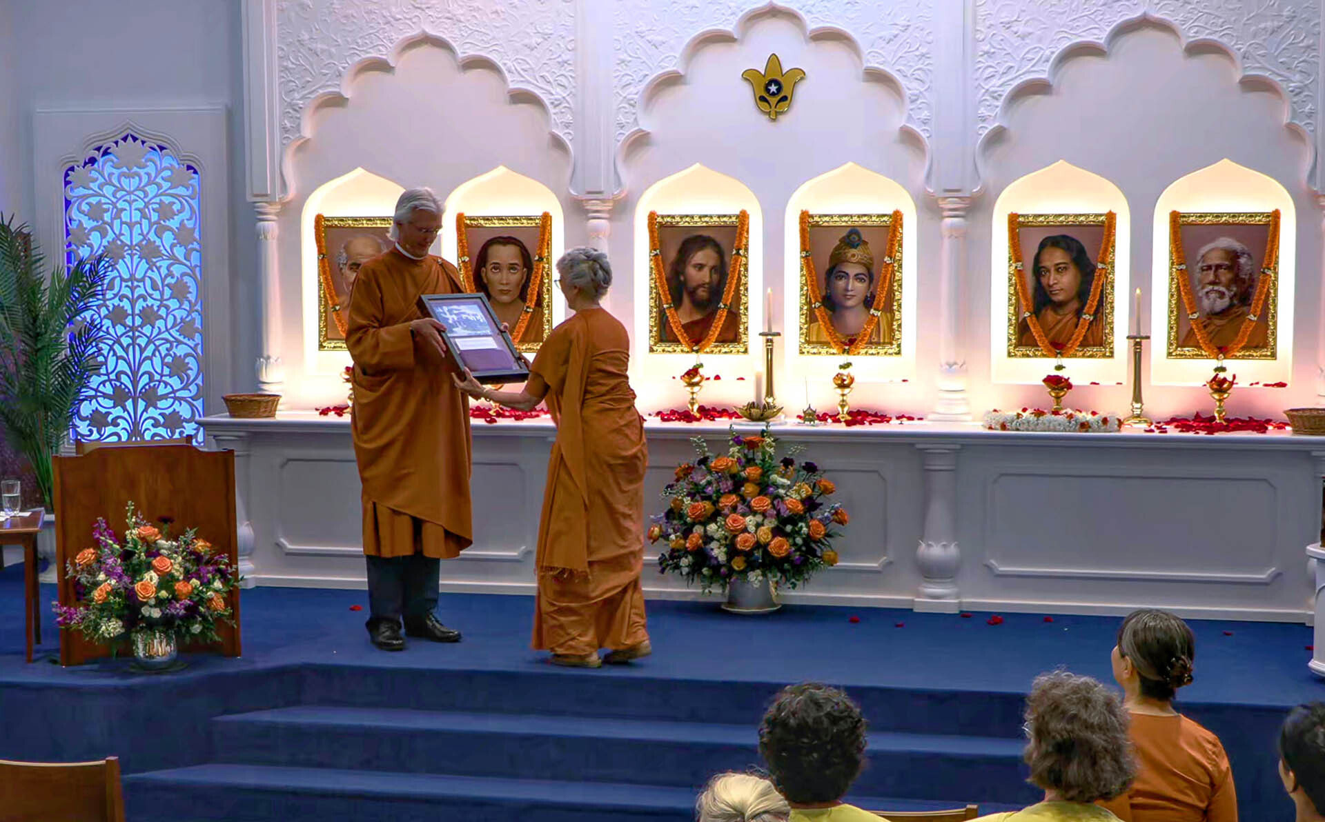 Brother Chidananda hands Sister Brahmani a gift for the Greenfield Retreat to mark the occasion: a photograph of Paramahansa Yogananda in front of the SRF International Headquarters Building and a portion of an original foundation brick from that iconic building.